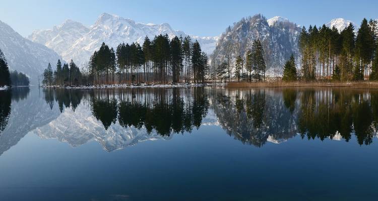 Spiegelruhiger Alpensee, der verschneite Gipfel und Kiefern an einem klaren Wintermorgen reflektiert.