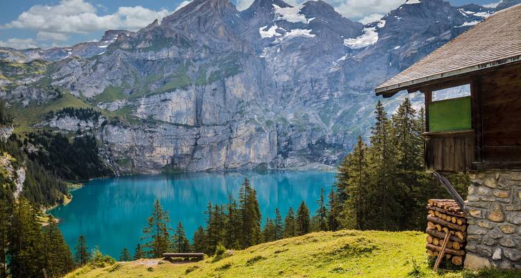Lago alpino turquesa vibrante acunado por acantilados dramáticos visto desde una cornisa cubierta de hierba con una cabaña rústica y troncos apilados.