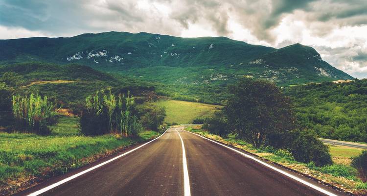 Carretera de asfalto recta que conduce hacia una cordillera verde bajo nubes dramáticas