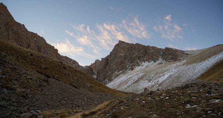 Paso de montaña rocoso y nevado al amanecer con nubes rosáceas y luna creciente arriba