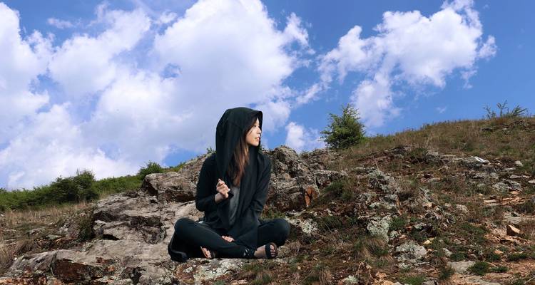 Mujer joven con atuendo negro con capucha sentada en una ladera rocosa bajo un cielo azul con nubes esponjosas