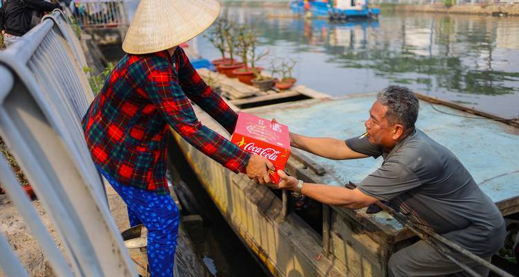 Hombre en barco recibiendo una caja de Coca-Cola de una mujer con sombrero cónico junto a la barandilla del río