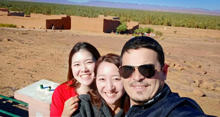Amigos sonrientes tomándose una selfie con un oasis de palmeras datileras y edificios de adobe al fondo.