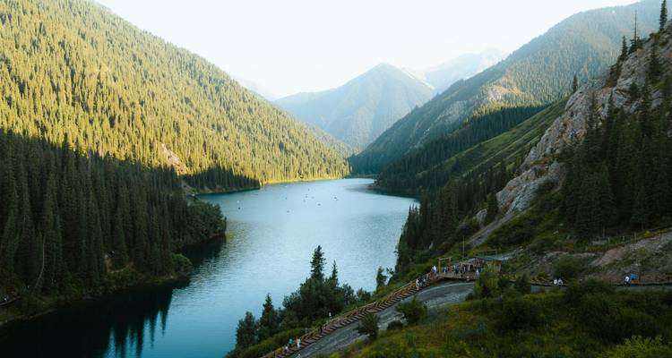 Un lac alpin pittoresque sinuant entre des pentes montagneuses densément boisées, avec des passerelles en bois et de minuscules bateaux visibles ci-dessous dans la lumière chaude de fin d'après-midi.