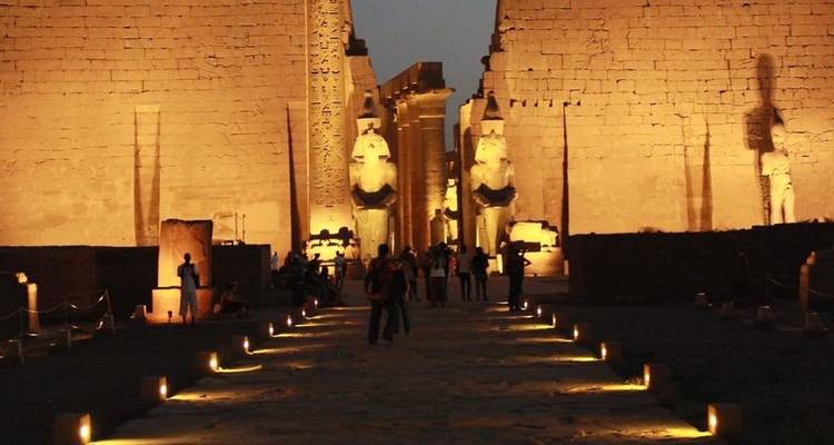Grote verlichte ingang van de Luxor Tempel bij schemering met bezoekers die tussen de kolossale standbeelden wandelen