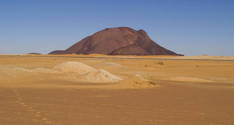 Isolierter dunkelbrauner Felsmonolith, der aus weitem goldenem Wüstensand unter klarem blauem Himmel aufragt.