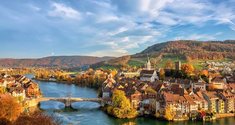 Pintoresco pueblo ribereño europeo con puente de piedra, follaje otoñal colorido y colinas.