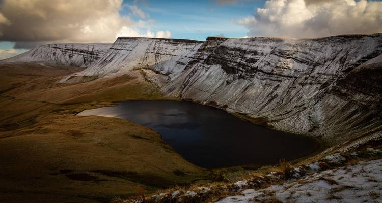 Dramatischer Hochlandsee, umschlossen von steilen schneegepuderten Klippen unter stimmungsvollen Himmeln