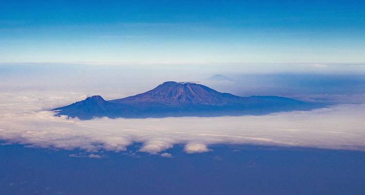 Le mont Kilimandjaro couronné de neige s'élève au-dessus d'une mer de nuages vue depuis les airs contre un ciel bleu dégagé.