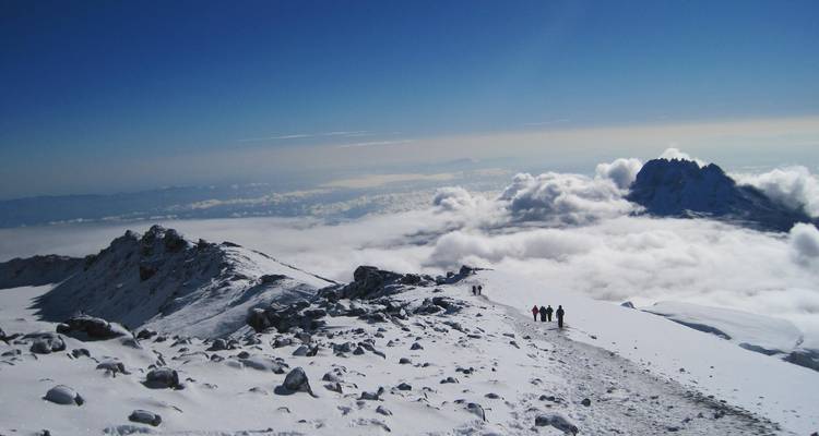 Des randonneurs marchent le long d'une crête enneigée en altitude sur le Kilimandjaro avec un vaste tapis de nuages en contrebas.