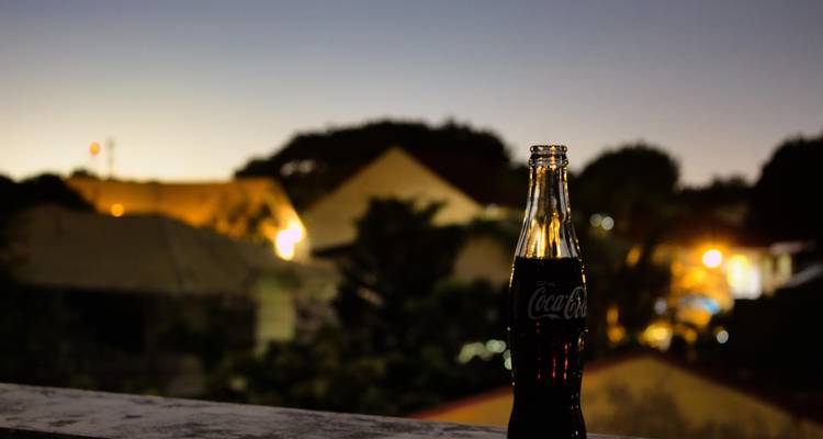 Coca-Cola Glasflasche auf Balkongeländer mit Abendlichtern und Dämmerhimmel.