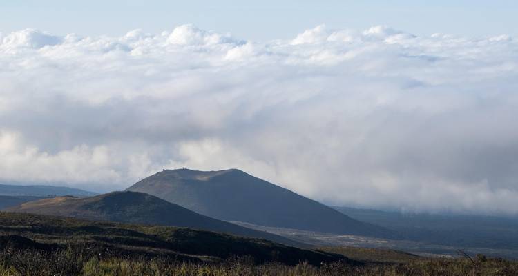 Cône volcanique bas émergeant de collines ondulantes sous un manteau de nuages.