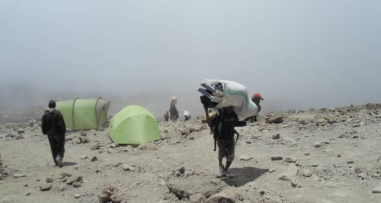 Des porteurs traversent une pente rocheuse et aride avec des tentes dressées au milieu du brouillard tourbillonnant de la montagne.