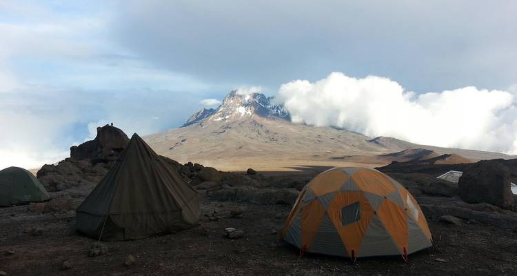 Tentes de camping plantées sur un terrain rocheux avec le mont Kilimandjaro couronné de nuages qui s'élève au-delà.