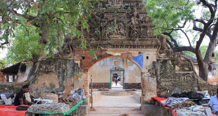 Oude stenen boog van een vervallen tempel met kleine marktkraampjes die schelpen en goederen verkopen onder schaduwrijke bomen