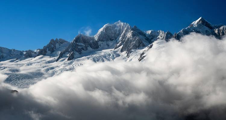 Des pics escarpés enneigés perçant au-dessus d'un épais manteau de nuages sous un ciel bleu profond.