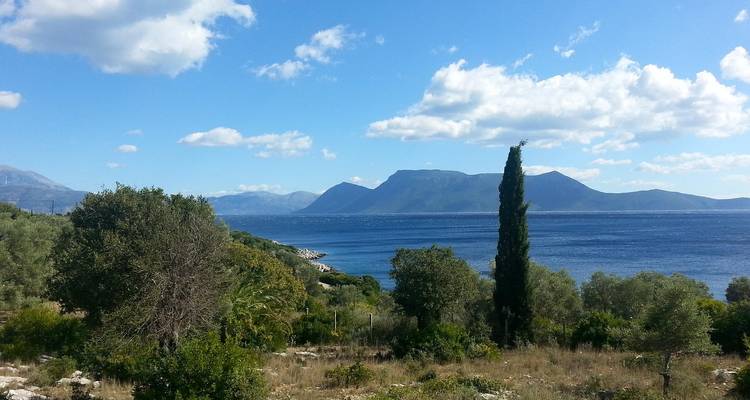 Mediterraans zeegezicht met cipressen, weelderige begroeiing en bergen aan de overkant van glitterend water.