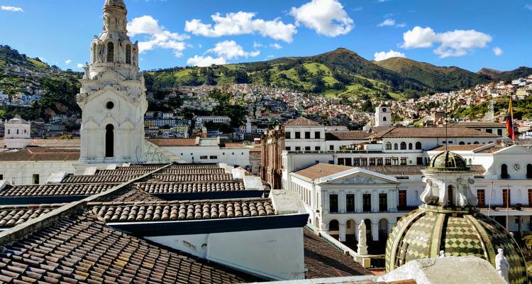 Rooftop view over Quito's historic center with white churches and green mountains under bright sky.