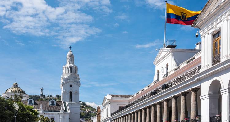 Government palace in Quito with Ecuadorian flag waving against blue sky, colonial church tower in background.