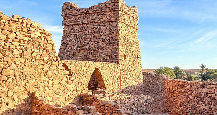 Tour de pierre ensoleillée et murs en ruine d'un ancien ksar dominent de clairsemées palmeraies.