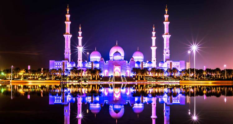 La Grande Mosquée Sheikh Zayed illuminée de teintes violettes vives se reflétant sur une eau calme la nuit.