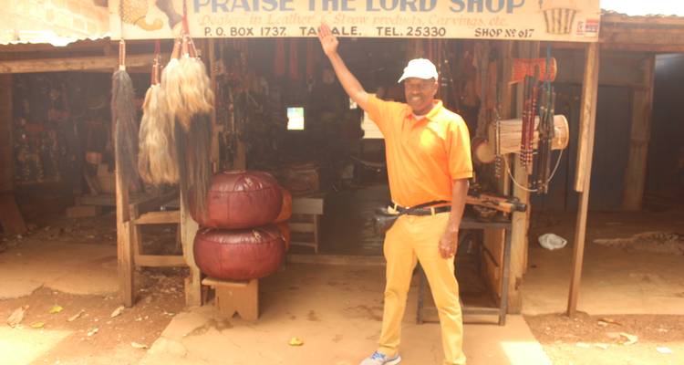 Man in orange outfit points at a signboard above a small handicraft shop on a dusty street.