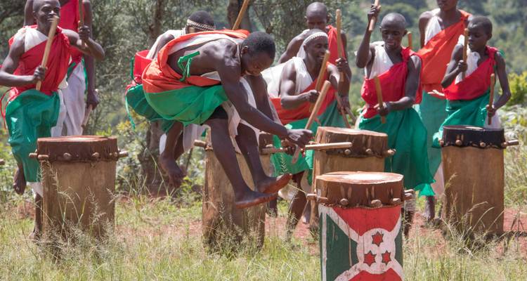 Burundische Trommler in leuchtend grünen und roten Kostümen treten energisch im Freien auf, während einer über eine Trommel springt.