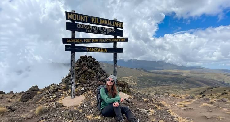 Un grimpeur est assis à côté du célèbre panneau de sommet sur un pic rocheux du mont Kilimandjaro au milieu de nuages tourbillonnants.