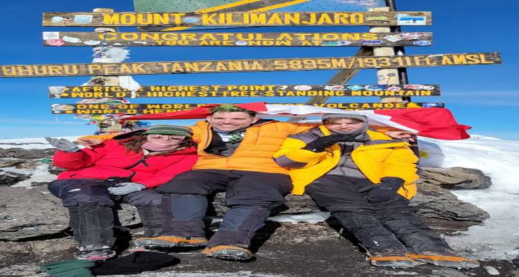 Trois alpinistes vêtus de tenues d'hiver célèbrent sous le panneau du sommet du Mont Kilimandjaro entourés de neige.