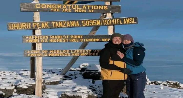 Couple en vestes d'hiver posant à côté du panneau sommital en bois au sommet enneigé du Kilimandjaro.