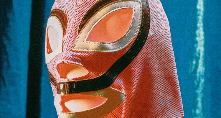 Een roze lucha libre masker tentoongesteld op een standaard tegen een groenblauw gordijn als achtergrond.