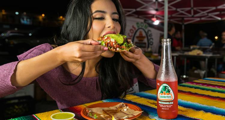 Een vrouw bijt enthousiast in een gevulde taco bij een kleurrijke buitenkraam 's nachts.