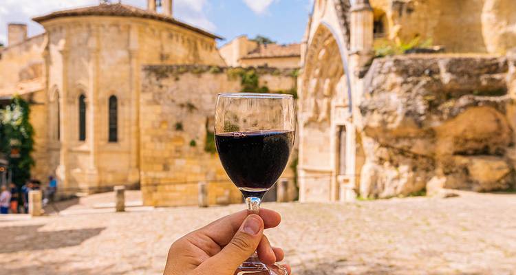 Une main tenant un verre de vin rouge devant un arrière-plan de bâtiments médiévaux en grès sous un soleil brillant.