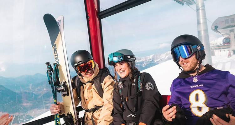 Trois jeunes skieurs portant des casques et des lunettes de ski qui sourient à l'intérieur d'un téléphérique au-dessus de montagnes enneigées