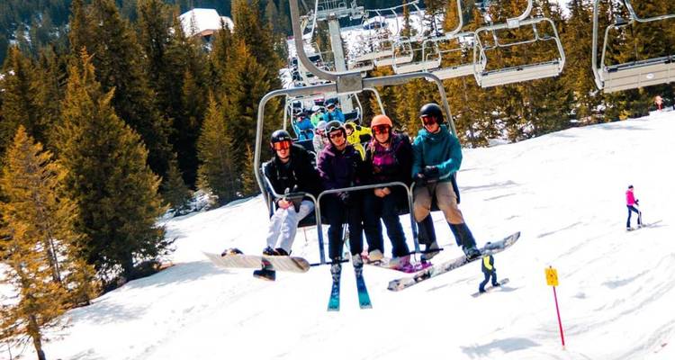 Groupe de snowboardeurs souriants sur un remontée-pente ascendante d'une pente alpine enneigée bordée de sapins.