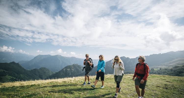 Vier Wanderer mit Rucksäcken gehen entlang eines Alpenwiesenrückens mit dramatischen Bergspitzen und einem strahlend blauen Himmel hinter ihnen.