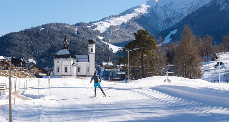 Ein einsamer Langläufer gleitet an einer malerischen Alpinekirche vorbei, mit bewaldeten Bergen und Skipisten im Hintergrund.