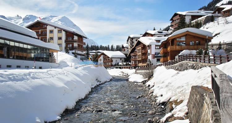 Ein schneebedecktes Alpendorf säumt einen engen Flusslauf unterhalb von schneegekrönten Gipfeln.