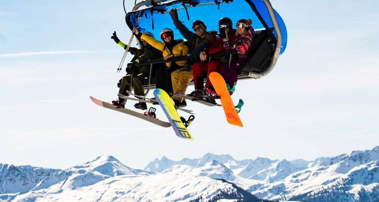 Vier Snowboarder jubeln von einem Sessellift hoch über verschneiten Gebirgsketten.