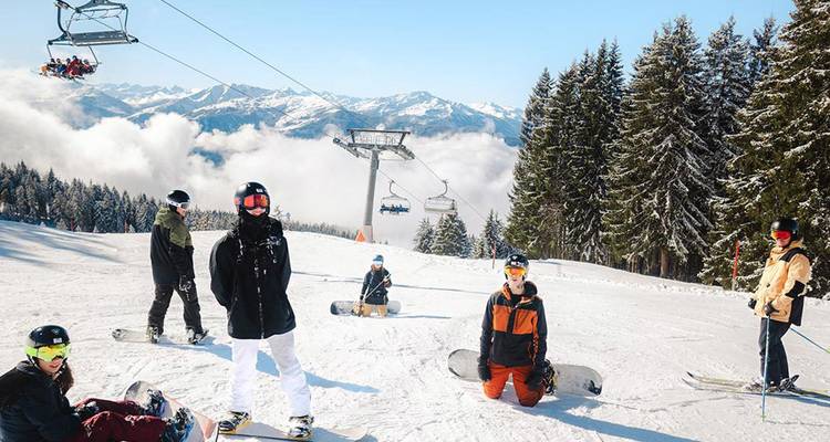 Snowboarders and skiers pause on a bright alpine slope with chairlifts and panoramic mountain views.