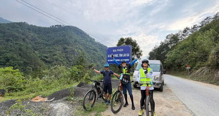 Cyclists celebrate beside a border sign on a mountain road with forested hills around.