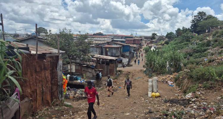 Residents walk along a rough dirt lane in a densely built informal settlement under cloudy skies.