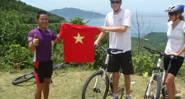 Cyclists hold a Vietnamese flag on a scenic hillside overlooking a coastal bay.