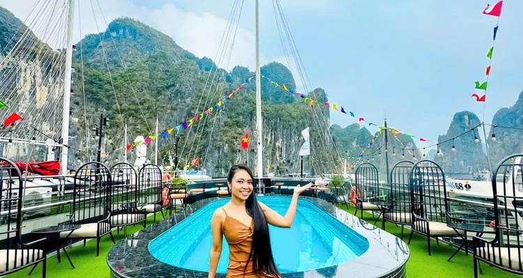 Een vrouw poseert op het dek van een jacht in Halong Bay met ruige kalksteen karstformaties en feestelijke vlaggen op de achtergrond.
