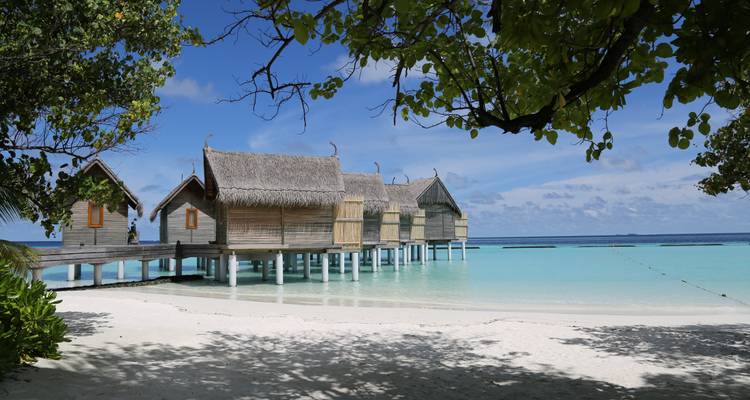 Reeks houten strandvilla's op palen boven een kalme, helder lagune omzoomd door wit zand