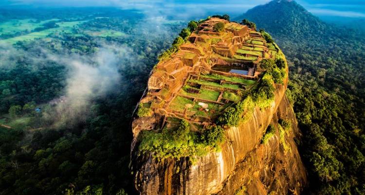 Dramatic aerial view of Sigiriya Lion Rock fortress rising above lush forest and morning mist.