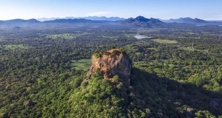 Wide aerial panorama of Sigiriya Rock rising from dense forest with distant mountain range.