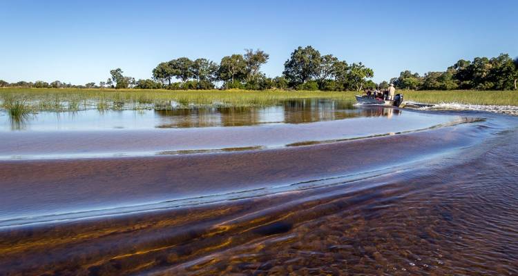 Motorboat speeding across a calm river creating ripples under a clear blue sky with grassy banks.