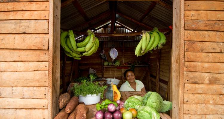 Een verkoper glimlacht vanaf een rustieke houten kraam die overloopt van vers tropisch fruit en hangende bananen.