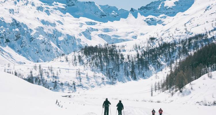Skifahrer durchqueren ein breites, schneebedecktes Tal mit dramatischen Alpengipfeln, die sich majestätisch darüber erheben.